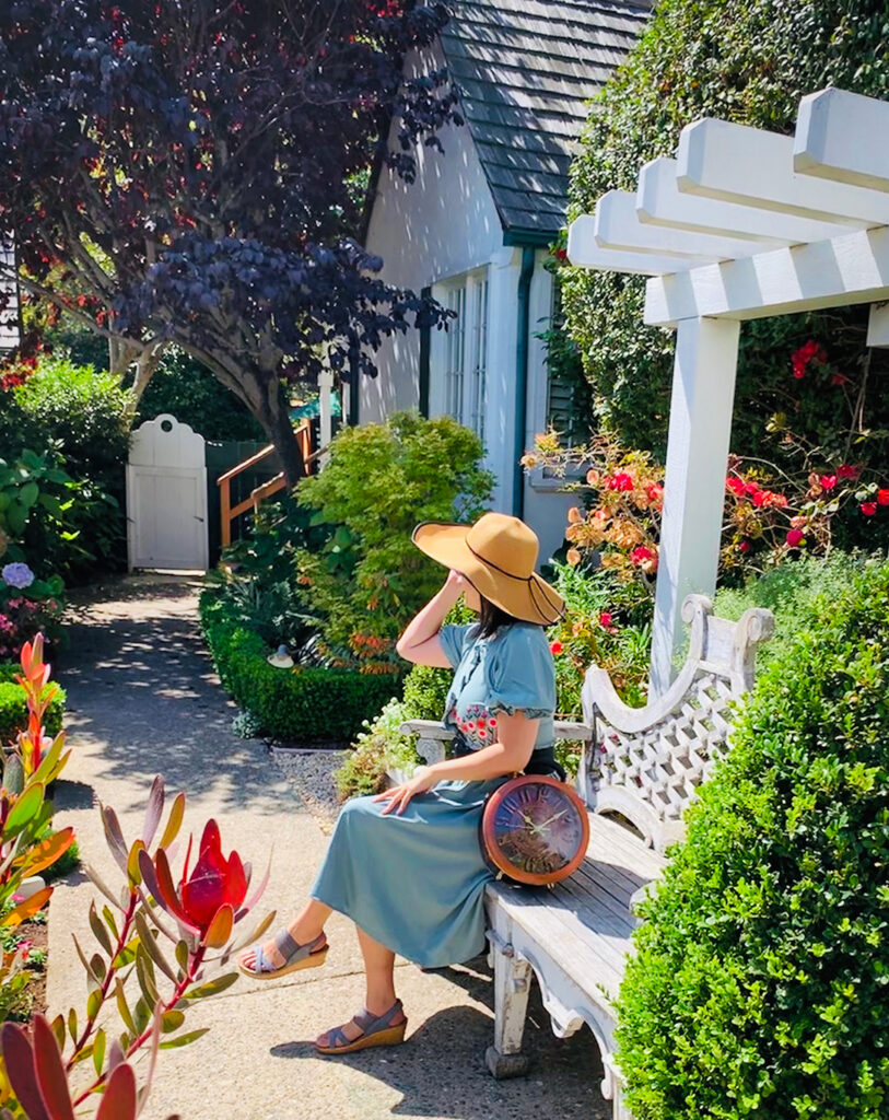 Girl enjoying sunny day in the gardens of a storybook Lincoln Green Inn in Carmel by the Sea, California.  Wearing a straw hat with a wonderland clock handbag.