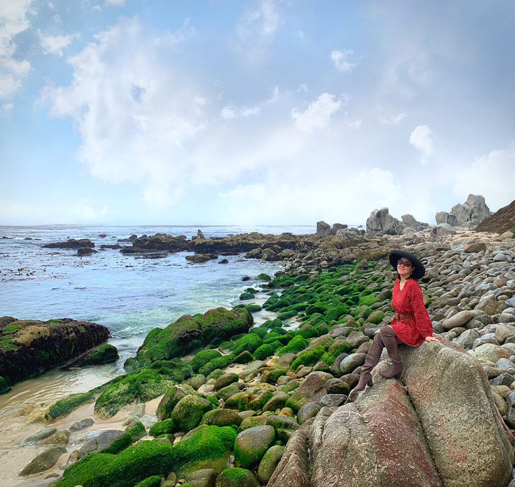 Hidden beach of mossy green rocks at Carmel Beach.  Girl in red dress and boots with black hat sitting on rocks.