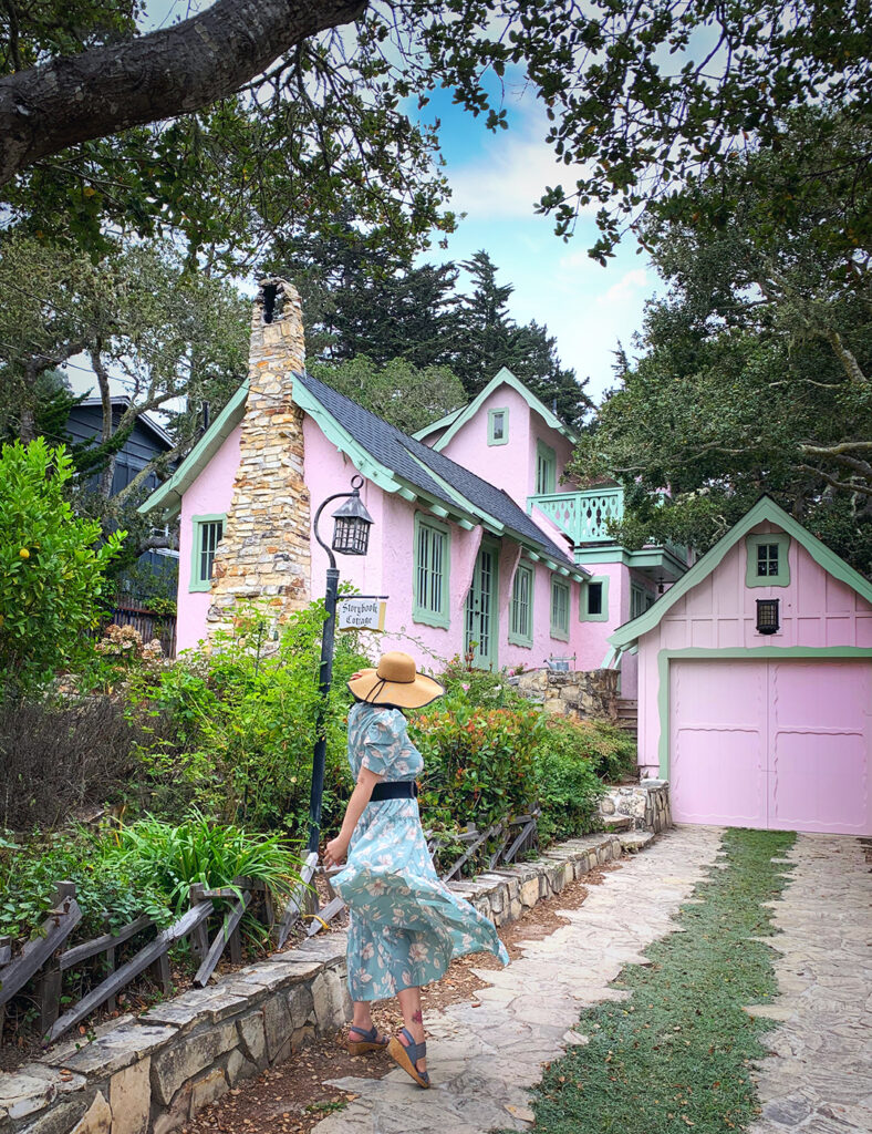 Cute pink gingerbread storybook fairy tale cottage in Carmel by the Sea designed by Hugh Comstock.  Girl in mint green flower dress with straw hat in front of cottage house.