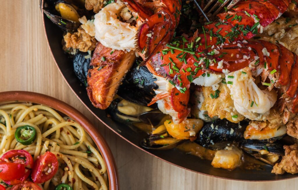 An overhead shot of pasta near a pan of fried lobster and meat with oysters on a wooden surface