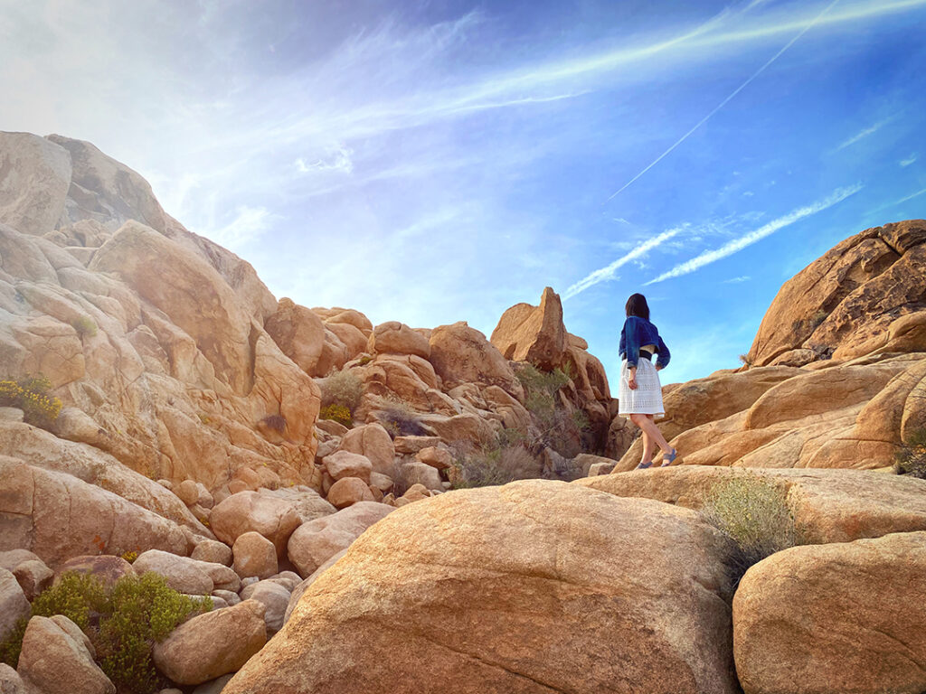 Girl in white dress and denim jacket standing high on pile of rocks in Joshua Tree National Park